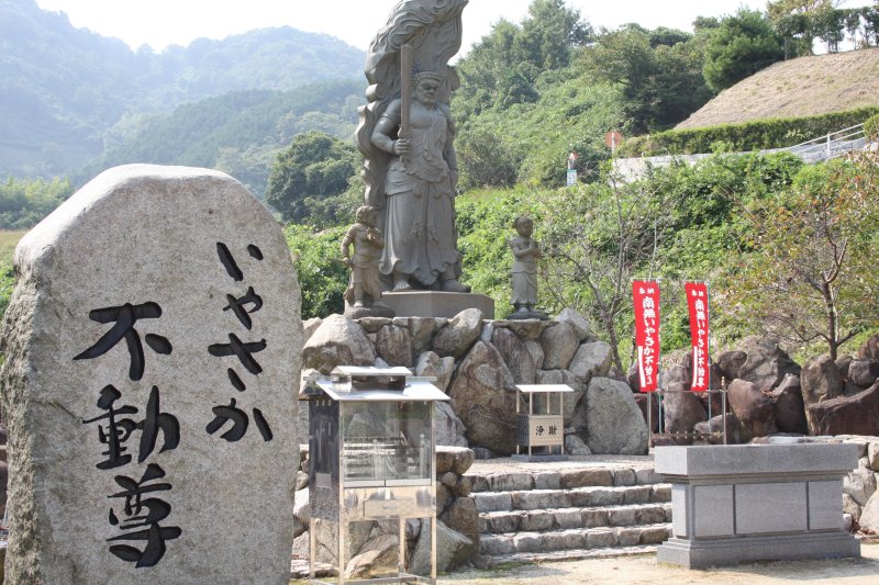 八坂寺の駐車場の一角には、ますます栄えるという意味を持つ「いやさか不動尊」が祀られている。背後には、奈良時代に法力を身につける修行が行われたと言われる大堂山が見える。